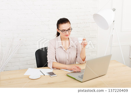 Middle aged businesswoman working in white modern office interior, with laptop and mobile cellphone. Female office work, woman in eye glasses, drinking coffee. High key, soft tone. woman in office. 137220409