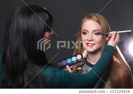 Backstage, fashion shooting. Make-up artist working with beauty model, applying eyeshadow palette, dark studio background Backstage, fashion shooting. Make-up artist working with beauty model, applying eyeshadow palette, dark studio background 137220428