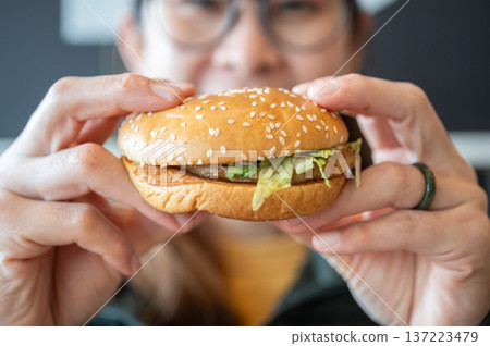 Cropped shot view of young Asian woman holding a hamburger in hands before eating. A hamburger is a sandwich consisting of a cooked meat patty on a bun or roll. Cropped shot view of young Asian woman holding a hamburger in hands before eating. A hamburger is a sandwich consisting of a cooked meat patty on a bun or roll. 137223479