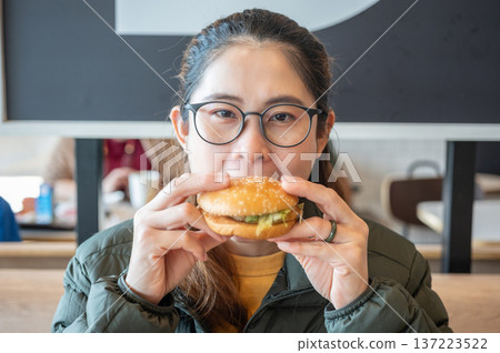 Cropped shot view of young Asian woman while eating a hamburger. A hamburger is a sandwich consisting of a cooked meat patty on a bun or roll. Cropped shot view of young Asian woman while eating a hamburger. A hamburger is a sandwich consisting of a cooked meat patty on a bun or roll. 137223522