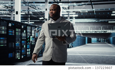 Data center system administrator tasked with reviewing code for AI automation scripts. African american man walking in server hub running diagnostics on equipment rigs, camera A 137223607