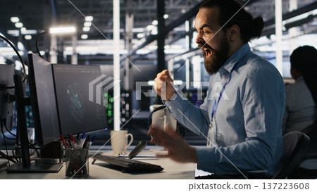 Server room IT specialist frustrated by difficult complex tasks in office. Stressed engineer developing neural network software while working extended hours in data center, camera A Server room IT specialist frustrated by difficult complex tasks in office. Stressed engineer developing neural network software while working extended hours in data center, camera A 137223608