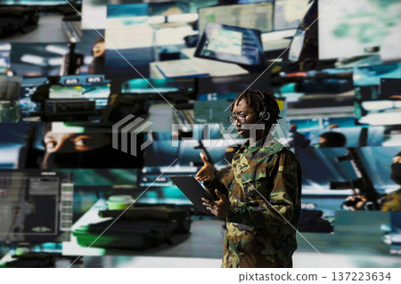 Armed forces operator manages battlefield communication networks from command hub using tablet. Soldier in command center provides real time combat support, talking in headset mic Armed forces operator manages battlefield communication networks from command hub using tablet. Soldier in command center provides real time combat support, talking in headset mic 137223634