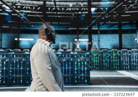 Engineer working in data center, listening music and overseeing infrastructure mainframes. Employee wearing audio headphones walking in server farm delivering web content to online customers 137223647