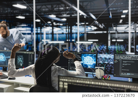 Exhausted data center female programmer stretching to ease back tension while using artificial intelligence. African american woman in server farm relieving pain after long hours at computer Exhausted data center female programmer stretching to ease back tension while using artificial intelligence. African american woman in server farm relieving pain after long hours at computer 137223670