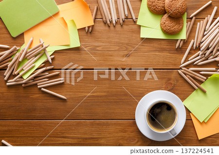 On a wooden table, a cup of coffee sits next to a plate of cookies. Colorful sticky notes and pencil sticks are scattered around, creating an inviting workspace atmosphere for creativity. 137224541