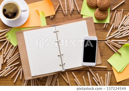A workspace is set up for creativity, featuring an empty binder, colorful sticky notes, a smartphone, a cup of coffee, and cookies scattered around, inviting productivity. A workspace is set up for creativity, featuring an empty binder, colorful sticky notes, a smartphone, a cup of coffee, and cookies scattered around, inviting productivity. 137224549