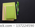 Three green notebooks of different sizes are stacked on a black surface alongside two green pens. The items create a minimalistic and organized workspace aesthetic, ideal for studying or planning. 137224590
