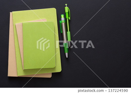 Three green notebooks of different sizes are stacked on a black surface alongside two green pens. The items create a minimalistic and organized workspace aesthetic, ideal for studying or planning. Three green notebooks of different sizes are stacked on a black surface alongside two green pens. The items create a minimalistic and organized workspace aesthetic, ideal for studying or planning. 137224590