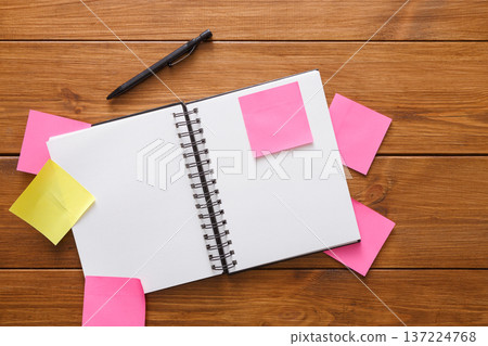 A notepad lies open on a wooden table. Several colorful sticky notes surround it, suggesting a brainstorming session. A pen rests beside the notepad ready for writing. 137224768