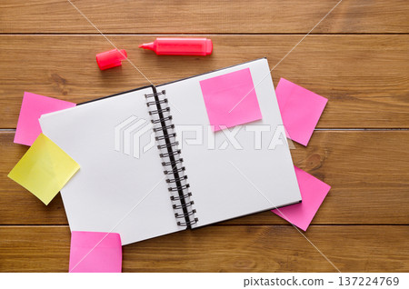 A blank spiral notebook sits on a wooden table surrounded by colorful sticky notes in pink and yellow. A pink highlighter is placed nearby, inviting creative brainstorming. 137224769