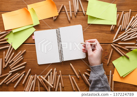 A person holds a pencil poised above an open notebook on a rustic wooden table. Surrounding the notebook are colorful sticky notes and scattered colored pencils, creating a vibrant workspace. A person holds a pencil poised above an open notebook on a rustic wooden table. Surrounding the notebook are colorful sticky notes and scattered colored pencils, creating a vibrant workspace. 137224789