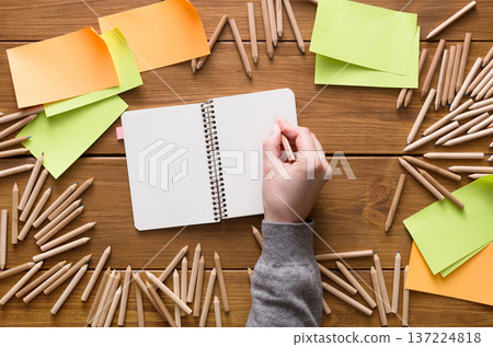 A hand reaches for a pencil above an empty notebook surrounded by colorful sticky notes and wooden pencils. The setting is a productive and creative workspace filled with ideas. 137224818