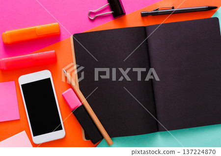 A neat workspace features various stationery items like highlighters, sticky notes, a smartphone, a pencil, and a blank notebook arranged on a vibrant, colorful surface. A neat workspace features various stationery items like highlighters, sticky notes, a smartphone, a pencil, and a blank notebook arranged on a vibrant, colorful surface. 137224870