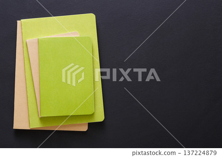 Three notebooks of varying sizes and shades of green are neatly stacked on a dark surface. The notebooks feature textured covers, creating a harmonious arrangement that highlights their colors. Three notebooks of varying sizes and shades of green are neatly stacked on a dark surface. The notebooks feature textured covers, creating a harmonious arrangement that highlights their colors. 137224879