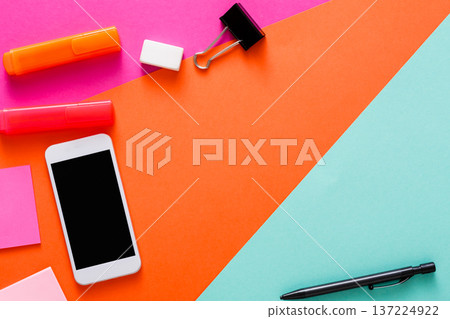 A vibrant and colorful workspace features a smartphone, sticky notes, a binder clip, and writing tools arranged neatly. The bright colors create an energetic atmosphere for creative work. 137224922