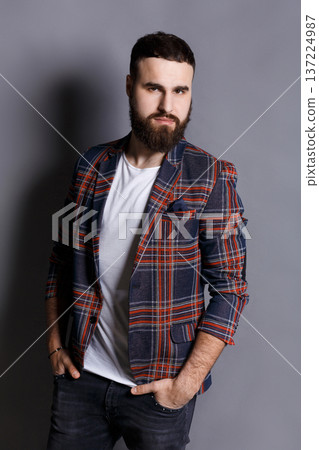 Handsome bearded man posing with hands in pocket. Portrait of young confident hipster in checkered jacket, looking at camera, gray studio background Handsome bearded man posing with hands in pocket. Portrait of young confident hipster in checkered jacket, looking at camera, gray studio background 137224987