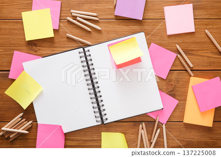 A notepad lies open on a wooden surface surrounded by vibrant sticky notes in various colors. Unused colored pencils are scattered nearby, suggesting an organized workspace for idea generation. 137225008