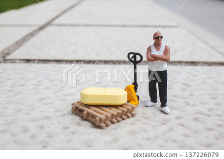 Miniature worker stands beside pallet jack holding yellow cargo Miniature worker stands beside pallet jack holding yellow cargo 137226079