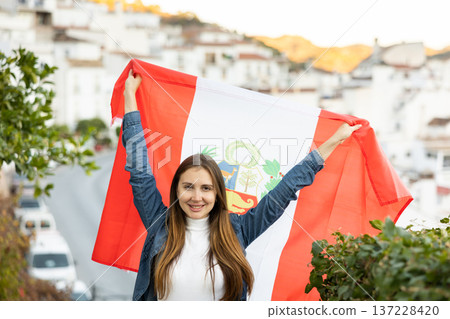 Excited young woman holding flag of Peru and smiling at camera, outdoors in town Excited young woman holding flag of Peru and smiling at camera, outdoors in town 137228420