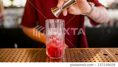 Preparing beverage. Alcohol mixing. Bartender pouring red liquid to glass with ice cubes. Measuring bright cocktail ingredient with jigger. Preparing beverage. Alcohol mixing. Bartender pouring red liquid to glass with ice cubes. Measuring bright cocktail ingredient with jigger. 137230029