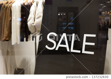 Retail window display highlights prominent SALE sign with selection of clothing items, including jackets and shirts, hanging on hangers, creating inviting shopping atmosphere Retail window display highlights prominent SALE sign with selection of clothing items, including jackets and shirts, hanging on hangers, creating inviting shopping atmosphere 137232630