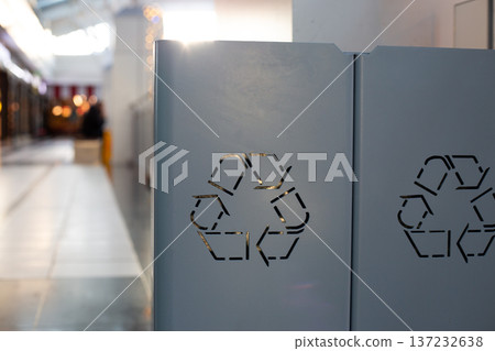 Recycling bin featuring cut out symbol positioned in contemporary indoor shopping area, with sunlight reflecting off its surface and clean, organized space visible in the background 137232638