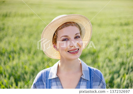 A smiling woman wearing a straw hat and plaid shirt stands in a lush green field on a sunny day A smiling woman wearing a straw hat and plaid shirt stands in a lush green field on a sunny day 137232697