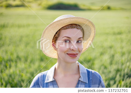 A smiling woman wearing a straw hat and plaid shirt stands in a lush green field under the warm sun 137232701