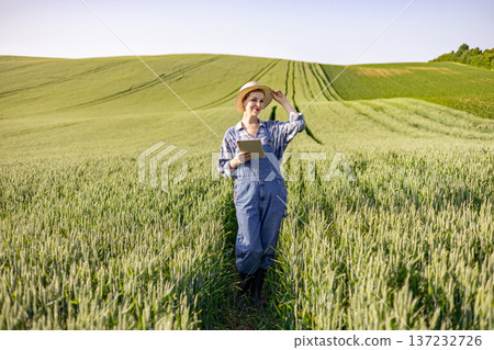 A young woman wearing overalls and a straw hat stands in a vast wheat field holding a tablet computer and smiling A young woman wearing overalls and a straw hat stands in a vast wheat field holding a tablet computer and smiling 137232726