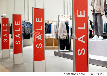 Interior of retail clothing store showcasing red sale signs with garments displayed on racks and shelves, creating vibrant shopping atmosphere Interior of retail clothing store showcasing red sale signs with garments displayed on racks and shelves, creating vibrant shopping atmosphere 137233452
