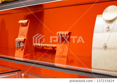 Earrings on orange stand displaying premium jewelry in store. Precious stones in earrings on jewelry shelf. Diamonds and gemstones for ear adornment. Shelf with premium jewelry 137233824