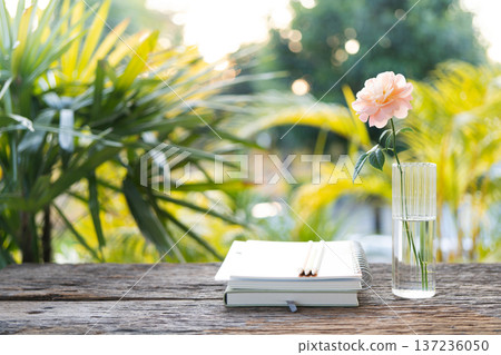 Stack of notebook and vase of rose on wooden table with backyard nature view Stack of notebook and vase of rose on wooden table with backyard nature view 137236050