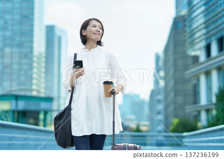 城市裡，一位女士提著行李箱，手裡拿著智慧型手機。 137236199