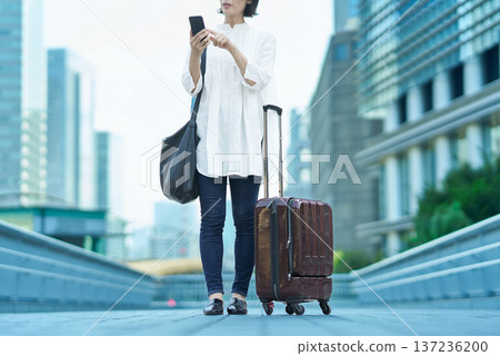 城市裡，一位女士提著行李箱，手裡拿著智慧型手機。 137236200