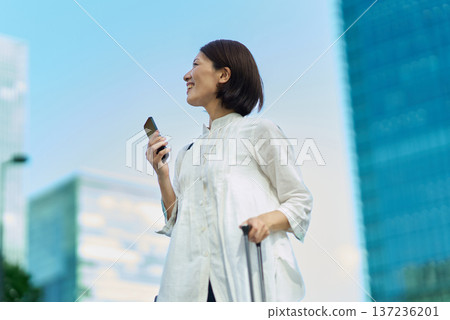 城市裡,一位女士提著行李箱,手裡拿著智慧型手機。 城市裡,一位女士提著行李箱,手裡拿著智慧型手機。 137236201