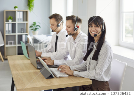 Smiling call center team with headsets working at laptops in bright office Smiling call center team with headsets working at laptops in bright office 137236421