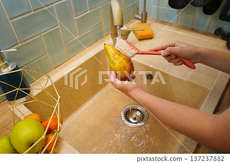 A vegan child practices kitchen hygiene, diligently brushing garden-fresh pear under running water in a sunny, domestic setting. 137237882