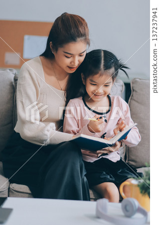 A cheerful Asian mother helps her young daughter do homework and color a book at home, while the father works on a laptop in the background. A cozy, bright, and modern family lifestyle. A cheerful Asian mother helps her young daughter do homework and color a book at home, while the father works on a laptop in the background. A cozy, bright, and modern family lifestyle. 137237941