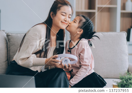 A cheerful Asian mother helps her young daughter do homework and color a book at home, while the father works on a laptop in the background. A cozy, bright, and modern family lifestyle. 137237955
