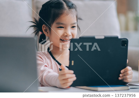 A cheerful Asian mother helps her young daughter do homework and color a book at home, while the father works on a laptop in the background. A cozy, bright, and modern family lifestyle. A cheerful Asian mother helps her young daughter do homework and color a book at home, while the father works on a laptop in the background. A cozy, bright, and modern family lifestyle. 137237956
