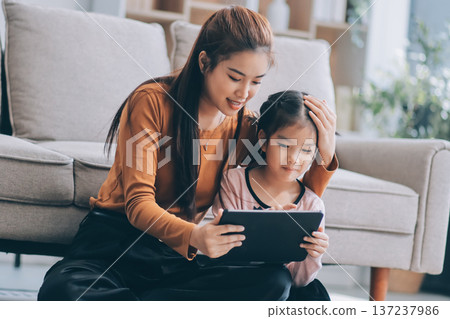 A cheerful Asian mother helps her young daughter do homework and color a book at home, while the father works on a laptop in the background. A cozy, bright, and modern family lifestyle. A cheerful Asian mother helps her young daughter do homework and color a book at home, while the father works on a laptop in the background. A cozy, bright, and modern family lifestyle. 137237986