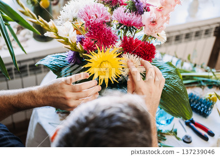 Hands of man arrange vibrant yellow and pink flowers in large white urn. Color therapy, vibrant floral palette, mood-enhancing decor, psychological impact of color, floral aesthetics. Hands of man arrange vibrant yellow and pink flowers in large white urn. Color therapy, vibrant floral palette, mood-enhancing decor, psychological impact of color, floral aesthetics. 137239046