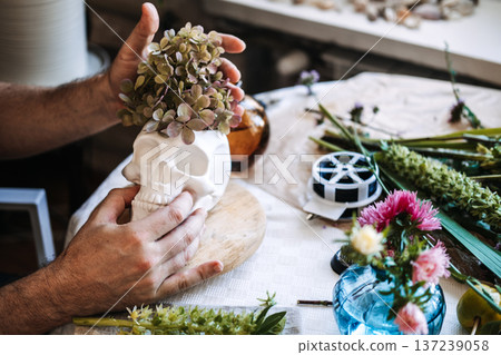 Hands place dried flowers into skull planter on table with floral tools. Moody home decor, gothic revival, maximalist interior design. Hands place dried flowers into skull planter on table with floral tools. Moody home decor, gothic revival, maximalist interior design. 137239058
