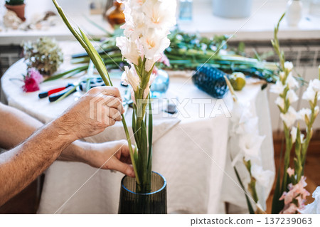 Hands of man carefully position tall gladiolus stems into fluted glass vase. Mental health wellness, DIY flower workshop, botanical therapy. Hands of man carefully position tall gladiolus stems into fluted glass vase. Mental health wellness, DIY flower workshop, botanical therapy. 137239063