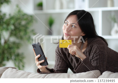 Pensive asian woman holding card and phone at home Pensive asian woman holding card and phone at home 137239495