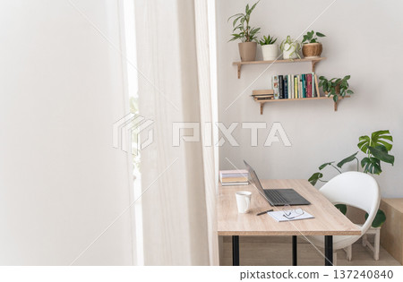 Modern home office workspace featuring laptop on wooden desk with white mug and notebook, surrounded by green plants bookshelves 137240840
