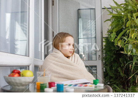 Young child in a warm fleece blanket relaxing on the balcony on cool spring day. Young girl at home. Young child in a warm fleece blanket relaxing on the balcony on cool spring day. Young girl at home. 137241337