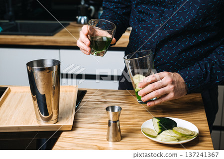 Hands hold glass of green liquid over mixing glass with cucumber slices on wooden counter. chlorophyll water benefits, functional green drinks, wellness mocktail trends, liquid chlorophyll recipes 137241367