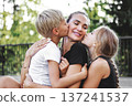 Beautiful Happy mother hugging two blonde children while kids kiss mom cheeks outdoors in summer park. Loving family moment, affection and joy, natural lifestyle and warm parent child relationship 137241537